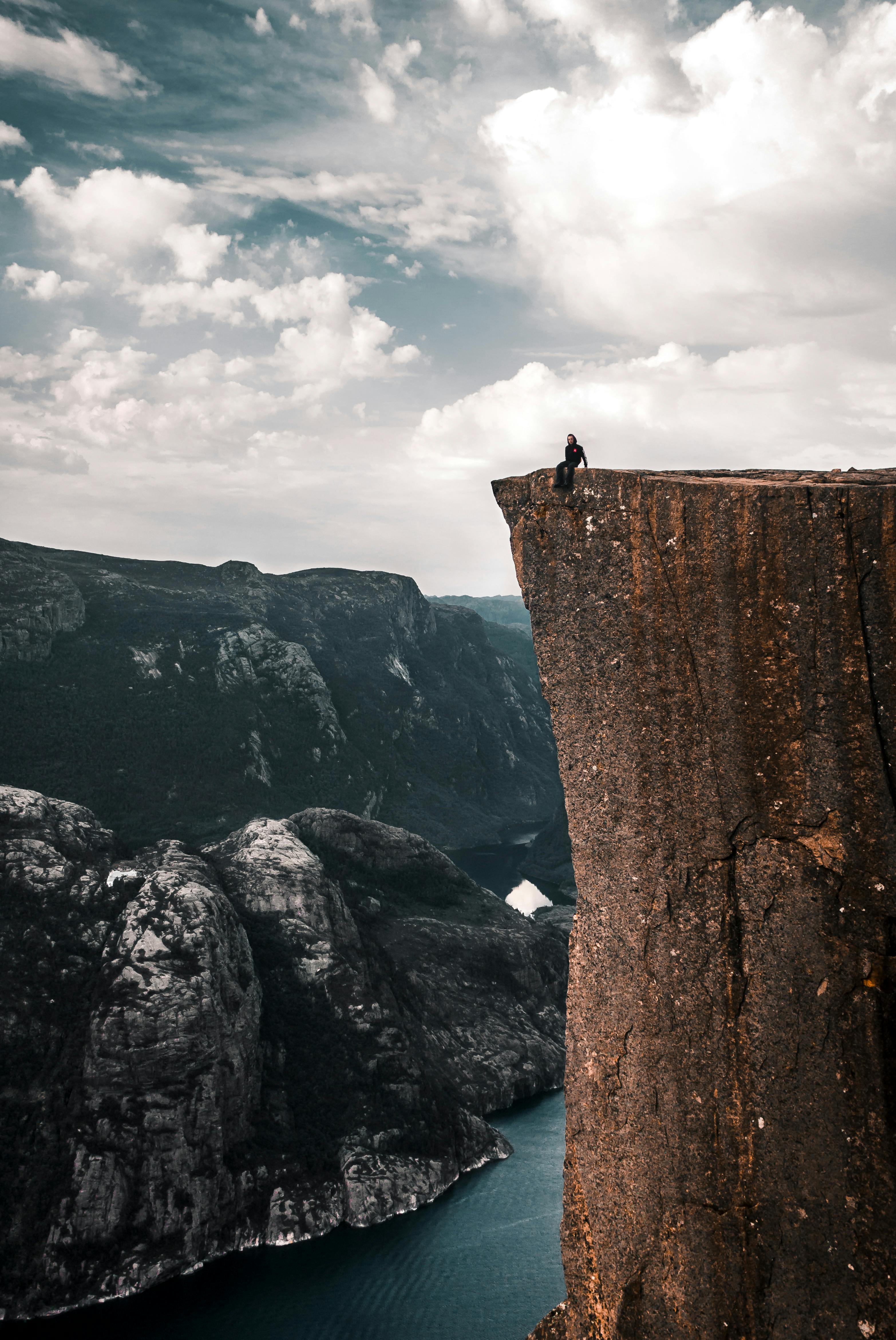 A lone figure seated at the edge of a towering cliff overlooking a fjord